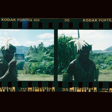 Kodak film scan of two images of a man from Vanuatu in traditional feathered headgear and natural clothing. In the 1st image he is holding a polaroid photo of people. In the 2nd he is smiling to camera. Palm trees and mountains in the background.