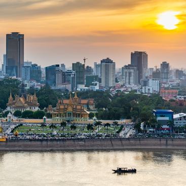 Rooftop view, looking across to the Riverside area of Cambodia's capital city. Sunset over Royal Palace and high rise buildings beyond, as a small boat drifts by towards the Mekong river.