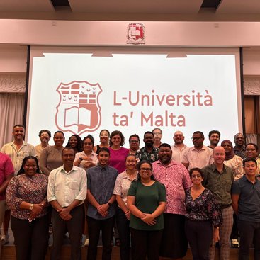 A cohort of professionals stand smiling in front of a projector screen with the words 'University of Malta'