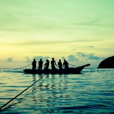 Six people silhouetted against the fading light, pulling fishing lines from a boat in the ocean