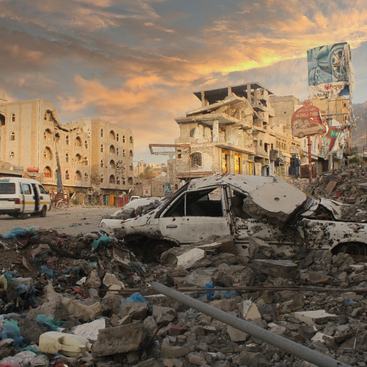 Taiz City, Yemen, a white car destroyed by bombings sits on a mass of rubble, destroyed buildings surround it both against the backdrop of mountains and a setting sun